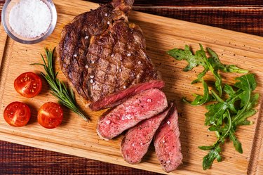 an overhead photo of sliced ribeye steak with arugula and tomatoes on a wooden cutting board